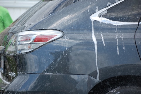 The rear side of a dark-colored vehicle is being washed, as evidenced by streams of white soap and water flowing down the car's surface. Reflections indicate a glossy finish, and part of a person wearing a green shirt is visible in the background.