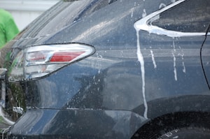 The rear side of a dark-colored vehicle is being washed, as evidenced by streams of white soap and water flowing down the car's surface. Reflections indicate a glossy finish, and part of a person wearing a green shirt is visible in the background.