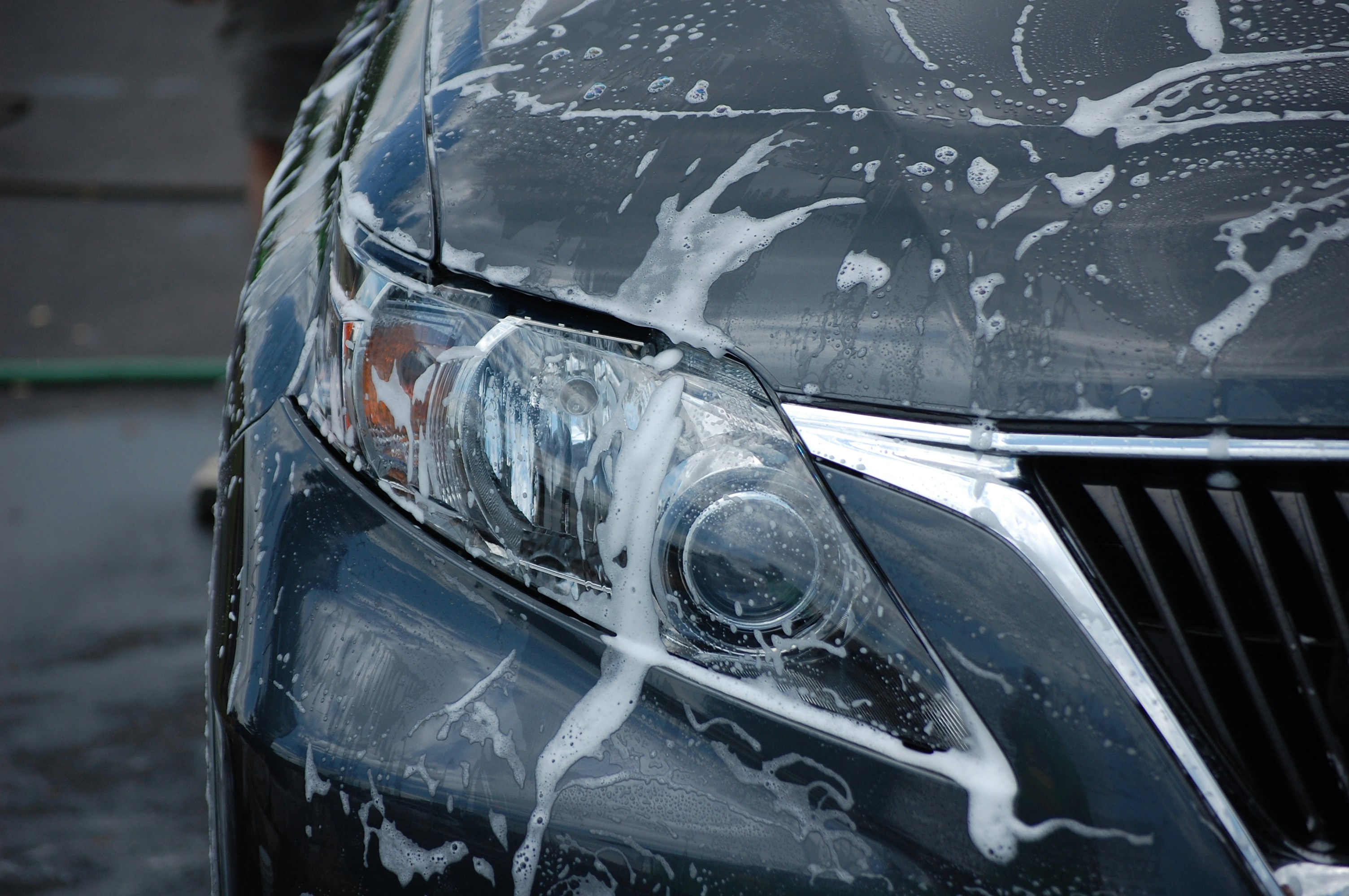 Car exterior being washed with foam, bright daylight, clean white suds on dark vehicle