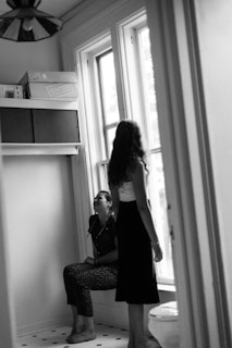 Two people are in a room with large windows. One person is sitting and laughing, while the other is standing, facing the window. The room has a shelf with boxes labeled Staples, and the light is soft, coming from the windows. The setting appears casual and candid.