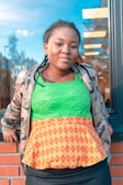 A young woman stands against a brick wall and in front of a large glass window. She has braided hair and is wearing a green lace top with a patterned orange peplum over a black skirt. She also wears a camouflage jacket and has her hands resting on the wall.