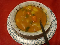 A bowl of chunky vegetable soup with visible pieces of carrot, potato, and green vegetables. It is served in a white bowl with decorative patterns, placed on a blue and white patterned plate. A spoon rests inside the bowl. The tablecloth is red.
