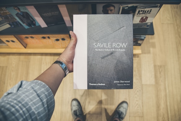 A person holds a book titled 'Savile Row: The Master Tailors of British Bespoke' by James Sherwood. The cover is sleek with a textured look, primarily gray with white text. The person is wearing a watch and casual shoes, standing on a wooden floor. In the background, there is a display shelf with other books.