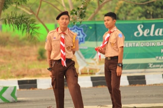 Two young individuals are wearing scout uniforms, including beige shirts and brown pants, with red and white striped scarves. They are standing outdoors near a road with signage in the background, suggesting a possible school or community event.