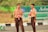 Two young individuals are wearing scout uniforms, including beige shirts and brown pants, with red and white striped scarves. They are standing outdoors near a road with signage in the background, suggesting a possible school or community event.