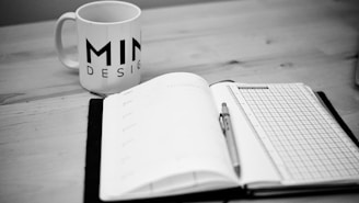 A black and white image of a notebook open on a desk, with a pen placed on top of it. To the left, there is a mug with the word 'MIND DESIGN' printed on it. The environment suggests a workspace or study area.