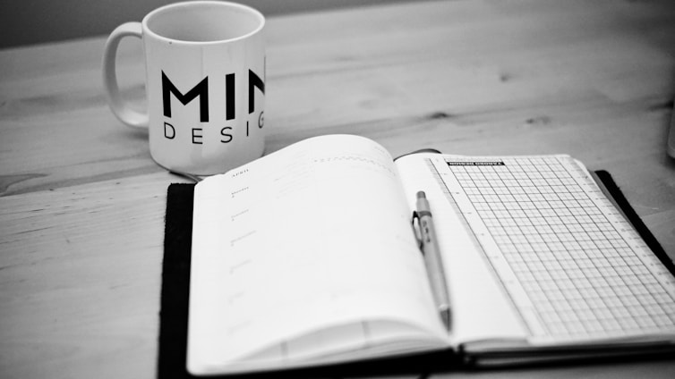 A black and white image of a notebook open on a desk, with a pen placed on top of it. To the left, there is a mug with the word 'MIND DESIGN' printed on it. The environment suggests a workspace or study area.