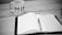 A black and white image of a notebook open on a desk, with a pen placed on top of it. To the left, there is a mug with the word 'MIND DESIGN' printed on it. The environment suggests a workspace or study area.