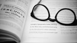 A pair of black glasses is placed on an open book, with the focus on printed text that includes various typographic styles and sizes. The layout suggests a design or typography manual, showcasing different font styles and layouts.