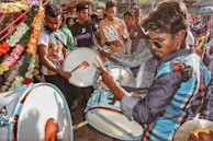 A group of people is engaged in playing drums in a vibrant setting with colorful flower decorations. The drummer in the foreground is wearing sunglasses, and there is a feeling of celebration with others clapping in the background.