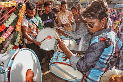 A group of people is engaged in playing drums in a vibrant setting with colorful flower decorations. The drummer in the foreground is wearing sunglasses, and there is a feeling of celebration with others clapping in the background.