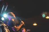 A person is holding a microphone, possibly singing or speaking on stage, illuminated by stage lights. The background is dark with circular light bokeh effects, highlighting the performer's presence.