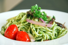 A plate of pasta coated with a green pesto sauce, garnished with sprigs of parsley, slices of red onion, and juicy cherry tomatoes. The dish appears fresh and vibrant, placed in a clean, white bowl.