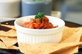 A white ceramic bowl filled with a vibrant salsa or vegetable dish is placed on a dark plate. The dish is garnished with fresh parsley and surrounded by several triangular pieces of flatbread or chips.
