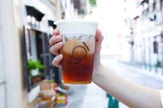 A hand is holding a clear plastic cup filled with a layered iced drink, featuring a milky foam layer on top and a brown tea at the bottom. The cup is branded with the logo 'KOI Thé'. The background is a blurred street scene with buildings and greenery.