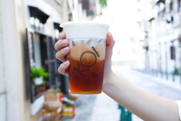 A hand is holding a clear plastic cup filled with a layered iced drink, featuring a milky foam layer on top and a brown tea at the bottom. The cup is branded with the logo 'KOI Thé'. The background is a blurred street scene with buildings and greenery.