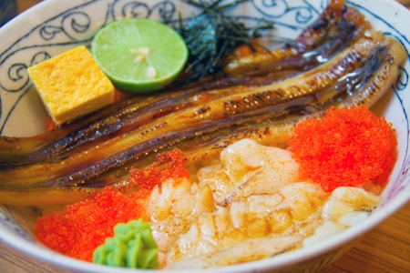 A bowl containing grilled eel, tamagoyaki (Japanese omelette), a slice of lime, seaweed, wasabi, and vibrant red fish roe on top of rice. The dish is presented in an intricately patterned bowl.
