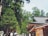 A vibrant scene features a traditional wooden building with a sloped roof, surrounded by lush green trees. Several people walk along the path, some carrying umbrellas for shade. The atmosphere is serene yet lively, suggesting a popular tourist destination or a cultural site.
