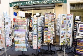 Outside a shop named 'Le Vieil Aix Cadeaux,' various racks display colorful postcards and greeting cards. The racks are filled with images of local landscapes, historical sites, and cultural art. A sign advertises a price for the items, and the shop's green awning is visible above.