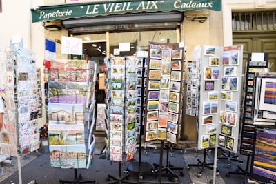 Outside a shop named 'Le Vieil Aix Cadeaux,' various racks display colorful postcards and greeting cards. The racks are filled with images of local landscapes, historical sites, and cultural art. A sign advertises a price for the items, and the shop's green awning is visible above.