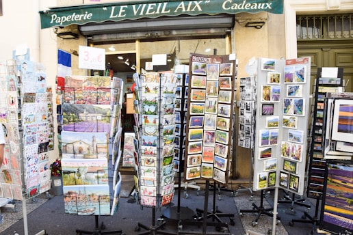 Outside a shop named 'Le Vieil Aix Cadeaux,' various racks display colorful postcards and greeting cards. The racks are filled with images of local landscapes, historical sites, and cultural art. A sign advertises a price for the items, and the shop's green awning is visible above.