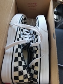 Pair of black and white checkered shoes inside a cardboard box with white laces and white midsoles. The box has some text on it that includes details and specifications.