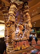 A large, detailed sculpture depicts a powerful figure in motion, appearing to be breaking two massive stone columns. The sculpture is set within a public space with people walking around. The name 'Samson' is prominently displayed at the base, suggesting a reference to the biblical story.