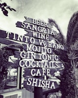 A wooden signpost features various beverage names in bold white letters. Surrounded by foliage, including a leafy plant and a palm tree, the backdrop includes a hint of a building with a thatched roof. The scene conveys a rustic, outdoor vibe.