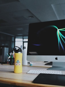 A workspace featuring a desktop computer with a bright, abstract screensaver on the monitor. In the foreground is a yellow water bottle with a sticker, placed on a wooden desk. A keyboard and stylus pad are also visible, with muted lighting suggesting an indoor setting.