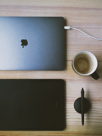 A workspace on a light wooden table with a closed silver laptop featuring a recognizable logo. A white charging cable is connected to the laptop. Beside it, there's a black graphics tablet with a stylus pen resting on a round pen holder. A black coffee mug with coffee stains inside is placed nearby.