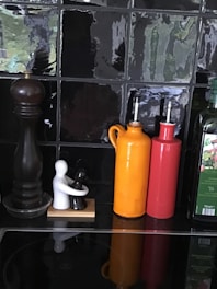 A kitchen countertop scene with a black pepper grinder, two ceramic figures in an embrace, and an orange and red oil dispenser, all set against a glossy black tiled backsplash. There is also a bottle of olive oil on the right.