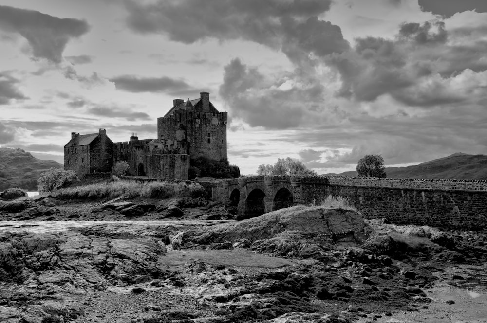 grayscale photography of castle beside body of water under cloudy sky