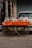 A street vendor setup with plastic bags filled with various grains and snacks displayed on a table covered with a vibrant orange cloth. The table is part of a cart with visible bicycle wheels, parked on a street in front of a closed shop shutter. The scene captures an urban marketplace atmosphere.