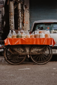 A street vendor setup with plastic bags filled with various grains and snacks displayed on a table covered with a vibrant orange cloth. The table is part of a cart with visible bicycle wheels, parked on a street in front of a closed shop shutter. The scene captures an urban marketplace atmosphere.