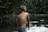 A child with short hair, wearing swim shorts, stands in a body of water with droplets splashing around. The background is lush with greenery, giving a natural and serene setting.