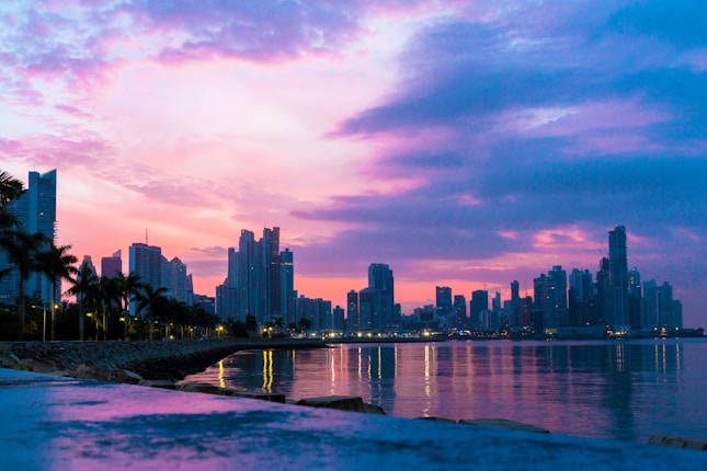 A scenic waterfront city skyline is silhouetted against a vibrant sunset with shades of pink, purple, and blue illuminating the sky. Tall skyscrapers line the horizon, and palm trees are visible along a stone-paved promenade. The calm water reflects the colorful sky and city lights.