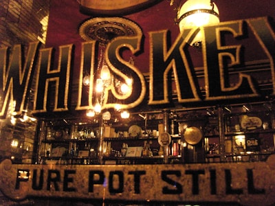 A dimly lit bar area with numerous bottles and glassware on shelves behind a large, bold sign displaying the word 'WHISKEY' and 'PURE POT STILL'. Ornate lighting fixtures and vintage decor contribute to a vintage ambiance.