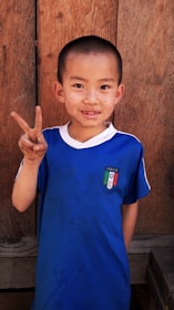 A young boy with short hair is standing in front of a wooden background and smiling. He is wearing a blue shirt with an Italian flag emblem and is holding up a peace sign with his right hand.
