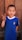 A young boy with short hair is standing in front of a wooden background and smiling. He is wearing a blue shirt with an Italian flag emblem and is holding up a peace sign with his right hand.