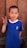 A young boy with short hair is standing in front of a wooden background and smiling. He is wearing a blue shirt with an Italian flag emblem and is holding up a peace sign with his right hand.