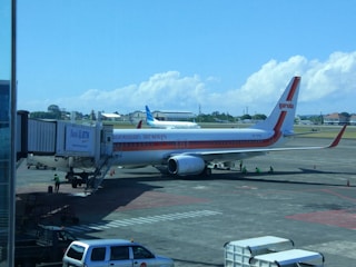 A Garuda Indonesia airplane is parked at an airport gate. The aircraft is connected to the jet bridge as ground staff members work around it. In the background, another plane can be seen on the tarmac against a backdrop of blue sky and scattered clouds.