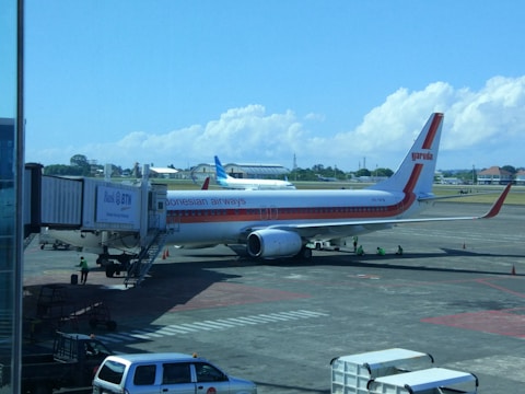 A Garuda Indonesia airplane is parked at an airport gate. The aircraft is connected to the jet bridge as ground staff members work around it. In the background, another plane can be seen on the tarmac against a backdrop of blue sky and scattered clouds.