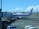 A Garuda Indonesia airplane is parked at an airport gate. The aircraft is connected to the jet bridge as ground staff members work around it. In the background, another plane can be seen on the tarmac against a backdrop of blue sky and scattered clouds.