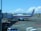 A Garuda Indonesia airplane is parked at an airport gate. The aircraft is connected to the jet bridge as ground staff members work around it. In the background, another plane can be seen on the tarmac against a backdrop of blue sky and scattered clouds.