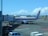 A Garuda Indonesia airplane is parked at an airport gate. The aircraft is connected to the jet bridge as ground staff members work around it. In the background, another plane can be seen on the tarmac against a backdrop of blue sky and scattered clouds.