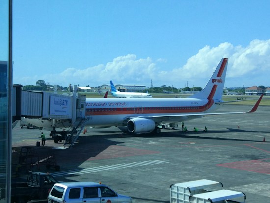 A Garuda Indonesia airplane is parked at an airport gate. The aircraft is connected to the jet bridge as ground staff members work around it. In the background, another plane can be seen on the tarmac against a backdrop of blue sky and scattered clouds.