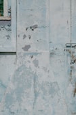 A section of a weathered wall with significant peeling paint and cracks. There is a window frame on the left side with some decay and a pipe structure on the right side.
