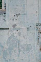 A section of a weathered wall with significant peeling paint and cracks. There is a window frame on the left side with some decay and a pipe structure on the right side.