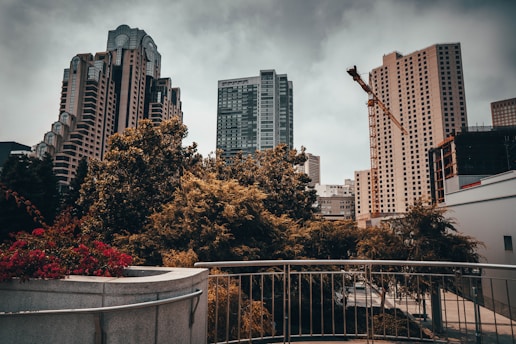 A cityscape featuring modern high-rise buildings with interesting architecture, including one with uniquely structured windows. In the foreground, lush green and reddish foliage can be seen, bordered by a metallic railing. A construction crane is visible in the background, indicating urban development.