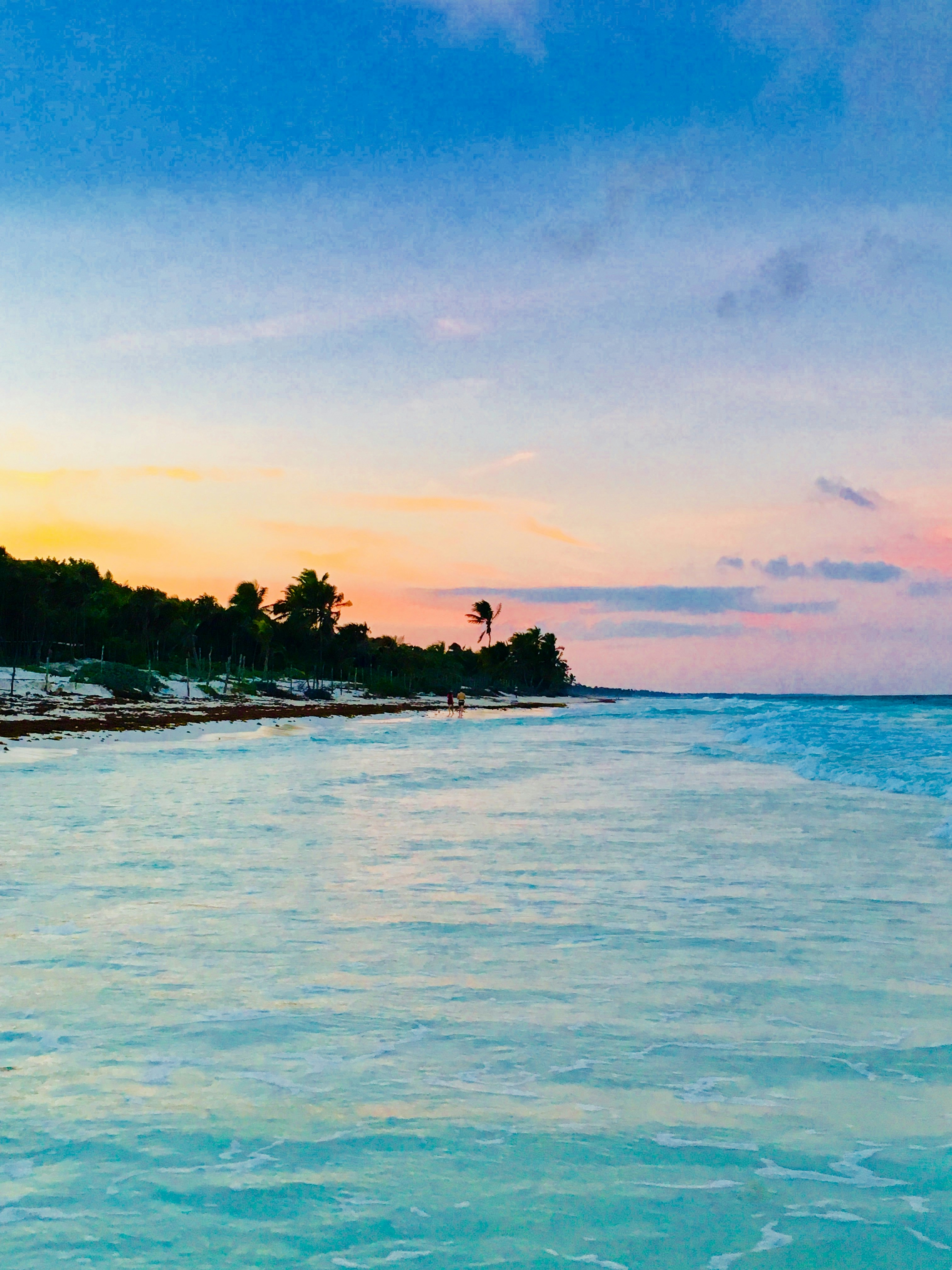 Couple walking on tropical beach at sunset, golden light reflecting on calm ocean water, romantic intimate atmosphere
